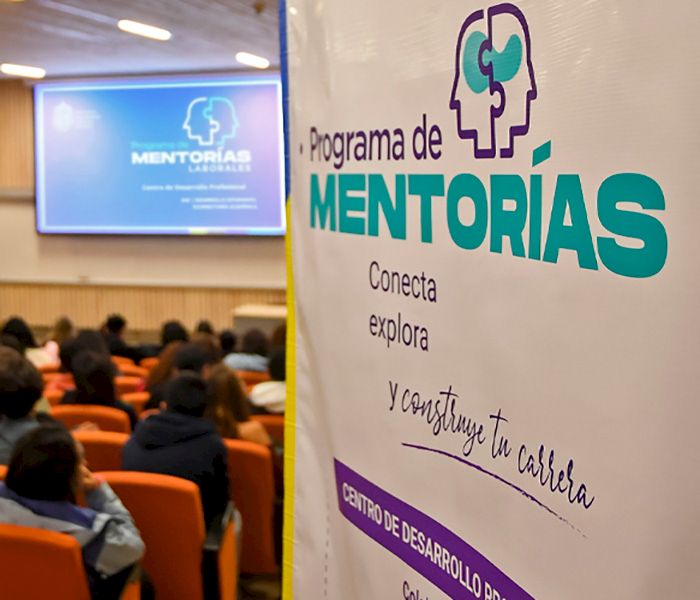 Pendón de programa de Mentorías junto a público de fondo.