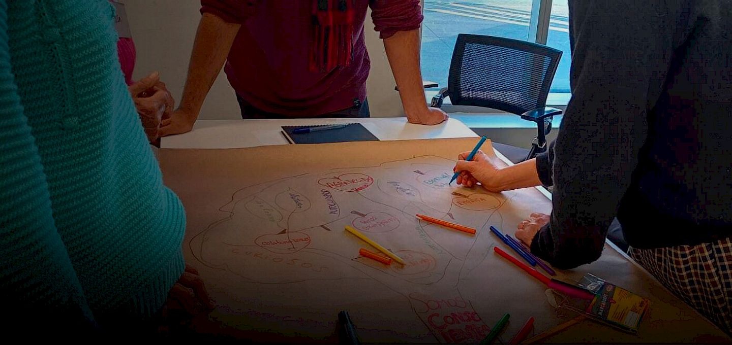 Personas en torno a un papelógrafo y lápices de colores, mientras una escribe sobre este.