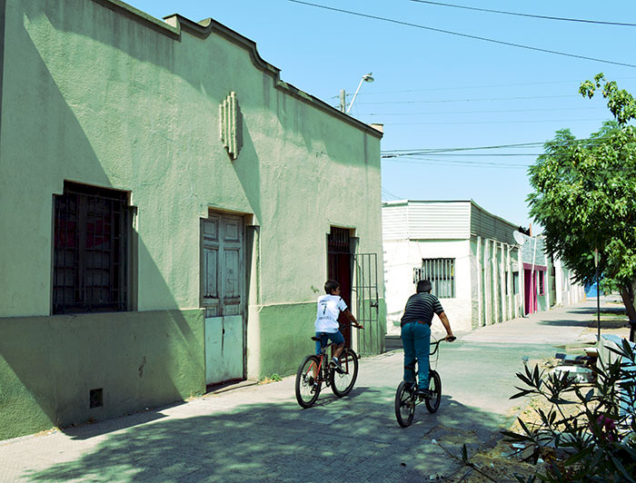 Niños andan en bicicleta por una vereda y se aprecia el frontis de una casa antigua.