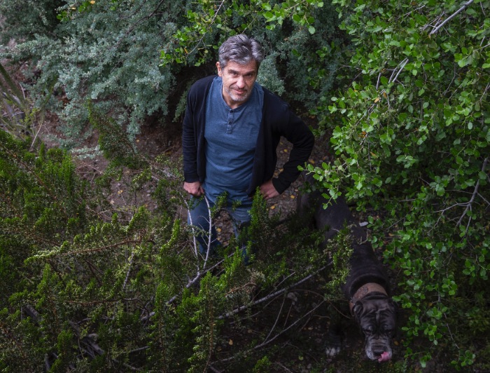 Mauricio Lima vistiendo popera, chaleco y jeans, en medio de árboles junto a un perro.