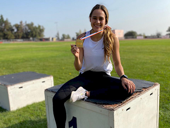 La la estudiante y velocista María Ignacia Montt muestra su medalla de oro sentada en el podio del primer lugar en una cancha de pasto.