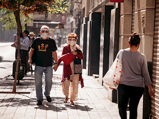 Pareja de adultos mayores con mascarilla y escudo facial caminando por una vereda de Santiago un día soleado.