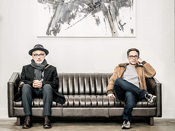 Elia Suleiman y  Gael García Bernal, sentados en un sofá de cuero café, mirando de frente a la cámara, a distancia y en silencio.