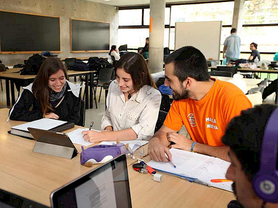 dos estudiantes mujeres y un estudiante hombre sentados alrededor de una mesa con lápices, cuadernos y tablet