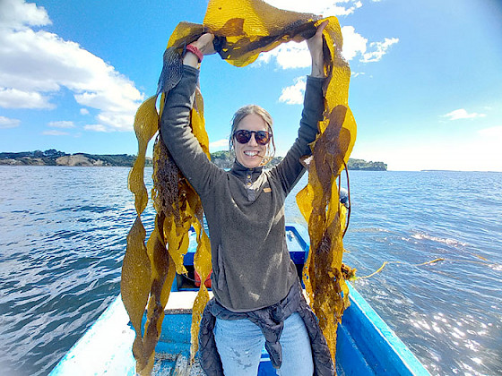 María José de la Fuente de pie sobre un bote levantando algas sobre su cabeza.
