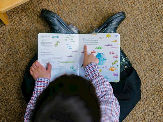 Niño sentado en el suelo con un libro abierto en su regazo, mostrando la lectura con un dedo.