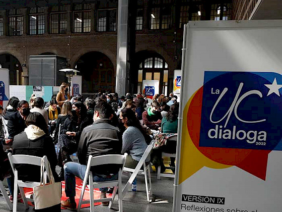 Personas sentadas en sillas conversando junto a un pendón de UC Dialoga.