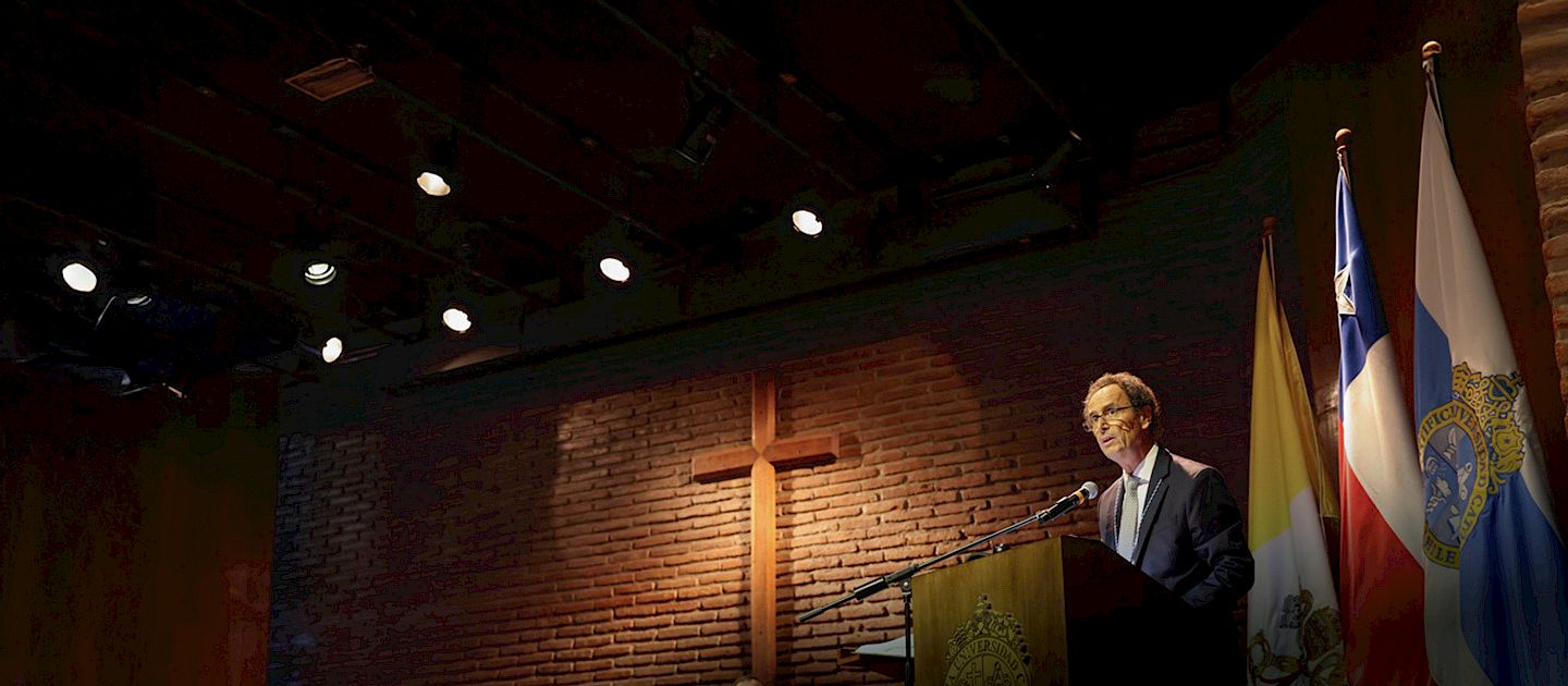 A man in a dark suit and tie speaks at a wooden podium in a dimly lit auditorium. A large wooden cross is mounted on a brick wall behind him. Flags, including those of Chile and the Vatican, are displayed on one side of the stage.