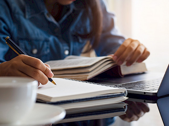 Mujer frente a un computador escribiendo en un cuaderno, junto a un libro y computador.