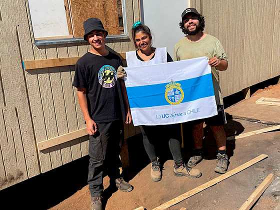 Voluntarios y habitantes damnificados frente a una vivienda sosteniendo bandera "La UC sirve a Chile"