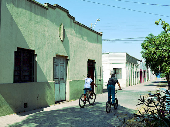 Niños andan en bicicleta por una vereda y se aprecia el frontis de una casa antigua.