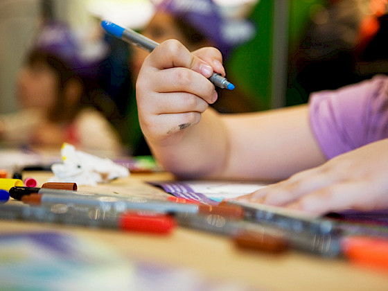 Mano de una niña sosteniendo un lápiz de color y un cuaderno, junto a lápices dispersos en una mesa.