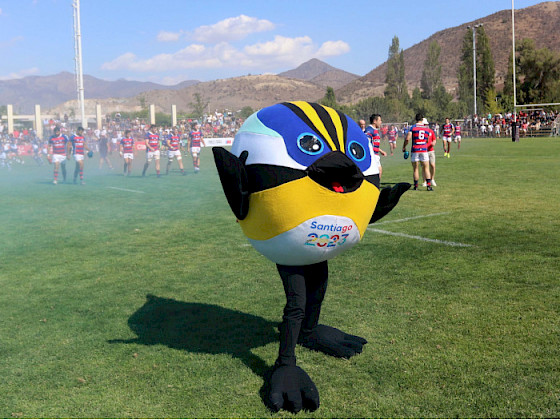 Mascota Fiu de los XIX Juegos Panamericanos y VII Parapanamericanos Santiago 2023, en una cancha con jugadores de rugby en el fondo.