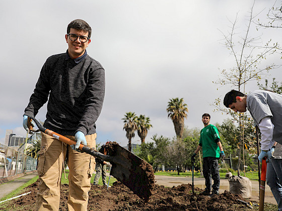 Estudiante sostiene una pala mientras participa de la forestación del campus San Joaquín, aparece un par de estudiantes más al fondo.