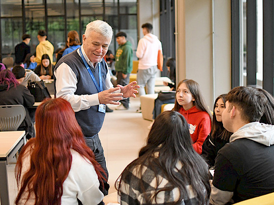 El director del programa Bachillerato Inicia conversando con un grupo de postulantes del programa en una sala; se ven otros jóvenes al fondo.