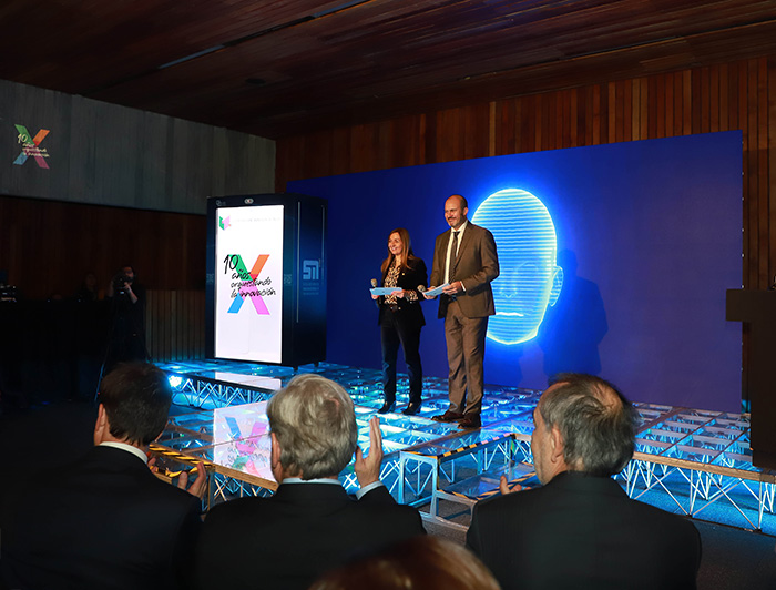 Los presentadores del evento, Marcela Briones y Ramón Molina, junto a un presentador virtual en una pantalla de fondo.