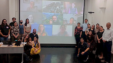 Grupo de participantes del Boot Camp de enero, en una sala de clases, junto a una pantalla con participantes online.