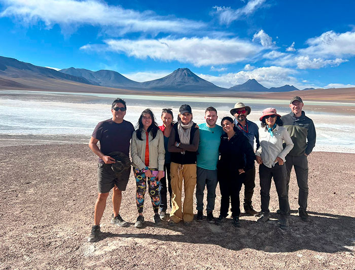 Grupo de investigadores que participa en el proyecto de estudio de la papa del desierto, en el desierto de Atacama, con un volcán y una laguna de fondo.