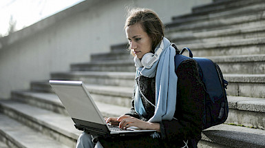 Mujer joven sentada en una escalera trabaja en su notebook, lleva unos audífonos y una mochila.
