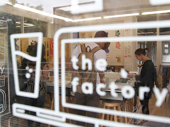 Personas trabajando en una sala tras un vidrio con la palabra The Factory