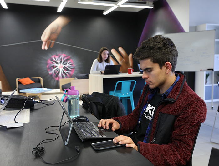 Un estudiante universitario trabaja en un computador, al fondo hay otra joven y tras ella un mural.