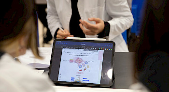 Una tablet muestra una figura de una mano humana, al rededor se ven siluetas de personas usando delantal blanco.