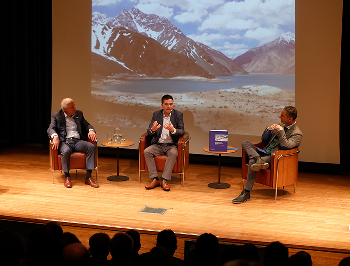 Presentación del libro: se ven los expositores sentados en el escenario junto a una foto de montañas y un lago de fondo.