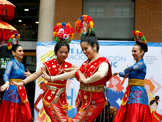 A group of women dressed in vibrant traditional attire perform a cultural dance. They wear red and gold outfits adorned with intricate patterns, along with colorful floral headpieces. The dancers move gracefully, smiling as they showcase their traditional choreography. In the background, there is a banner indicating an event at Pontificia Universidad Católica de Chile, celebrating a cultural festival.