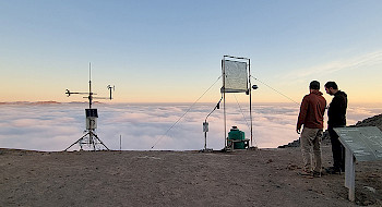 Dos jóvenes en terreno junto a instrumentos en un lugar árido y niebla de fondo