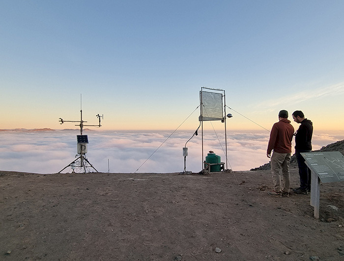 Dos jóvenes en terreno junto a instrumentos en un lugar árido y niebla de fondo