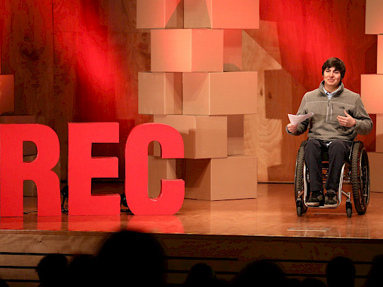 persona en silla de ruedas, sobre un escenario que tiene las letras R, E y C en rojo