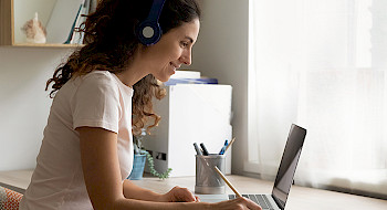 Mujer con audífonos sentada en un escritorio, escribiendo en un cuaderno mientras mira un computador portátil