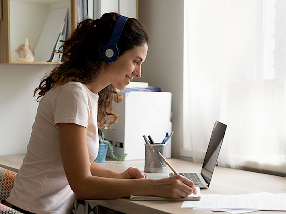 Mujer con audífonos sentada en un escritorio, escribiendo en un cuaderno mientras mira un computador portátil