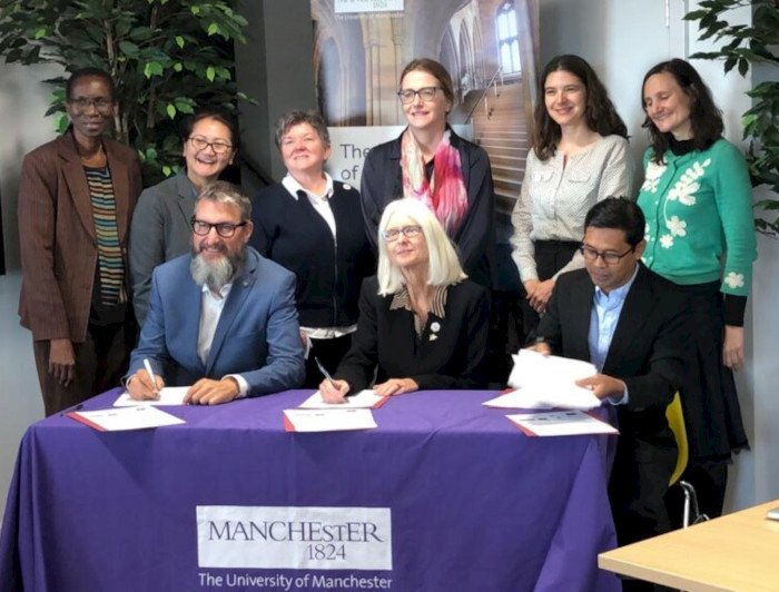 Group of representatives from universities signing an agreement at the University of Manchester, seated at a table with the Manchester logo.