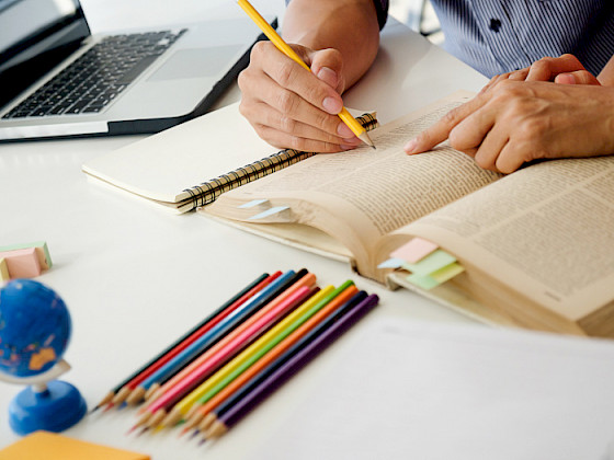 Close-up of hands writing in an open book on a desk, surrounded by colored pencils, a small globe, and a laptop in the background, symbolizing education and learning.