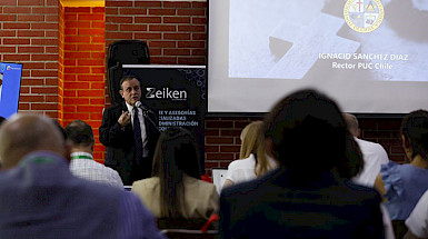 President Ignacio Sánchez, wearing a dark suit and tie, is giving a presentation in front of an audience in a conference room with exposed brick walls. He is standing next to a screen displaying a slide with his name.