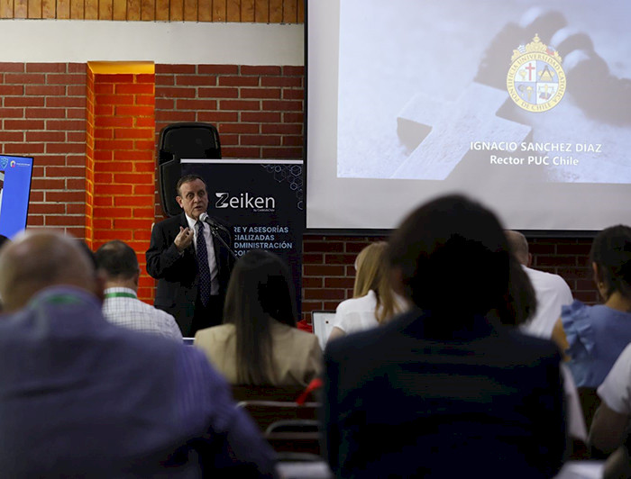 President Ignacio Sánchez, wearing a dark suit and tie, is giving a presentation in front of an audience in a conference room with exposed brick walls. He is standing next to a screen displaying a slide with his name.