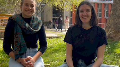 Two young women sitting on the grass at a university campus, smiling.