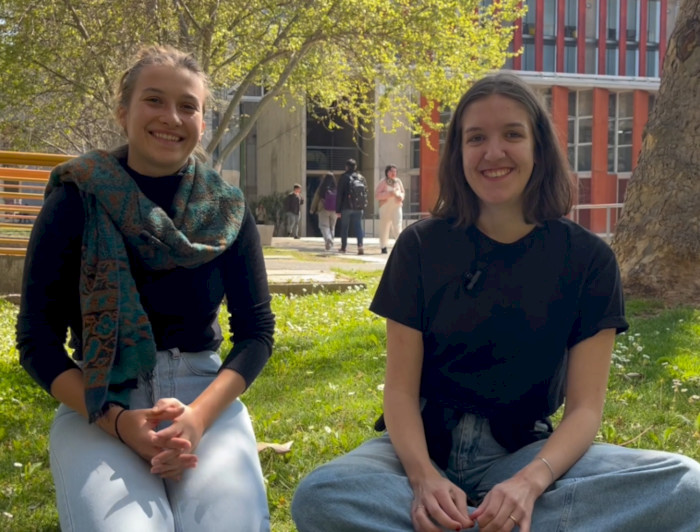 Two young women sitting on the grass at a university campus, smiling.