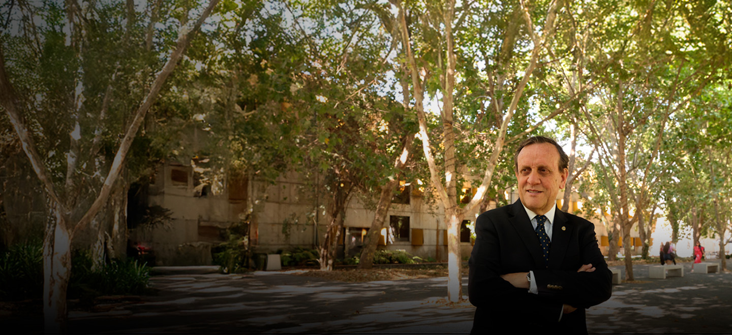 President Ignacio Sánchez stands with arms crossed in an outdoor setting at UC Chile, with a tree-lined walkway in the background.