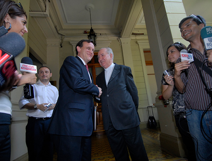 President Ignacio Sánchez shakes hands with Cardinal Francisco Javier Errázuriz in a formal setting, surrounded by journalists and media representatives.