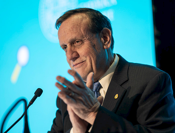 President Ignacio Sánchez delivers a speech at a formal event, standing at a podium with a microphone, against a blue-lit backdrop.