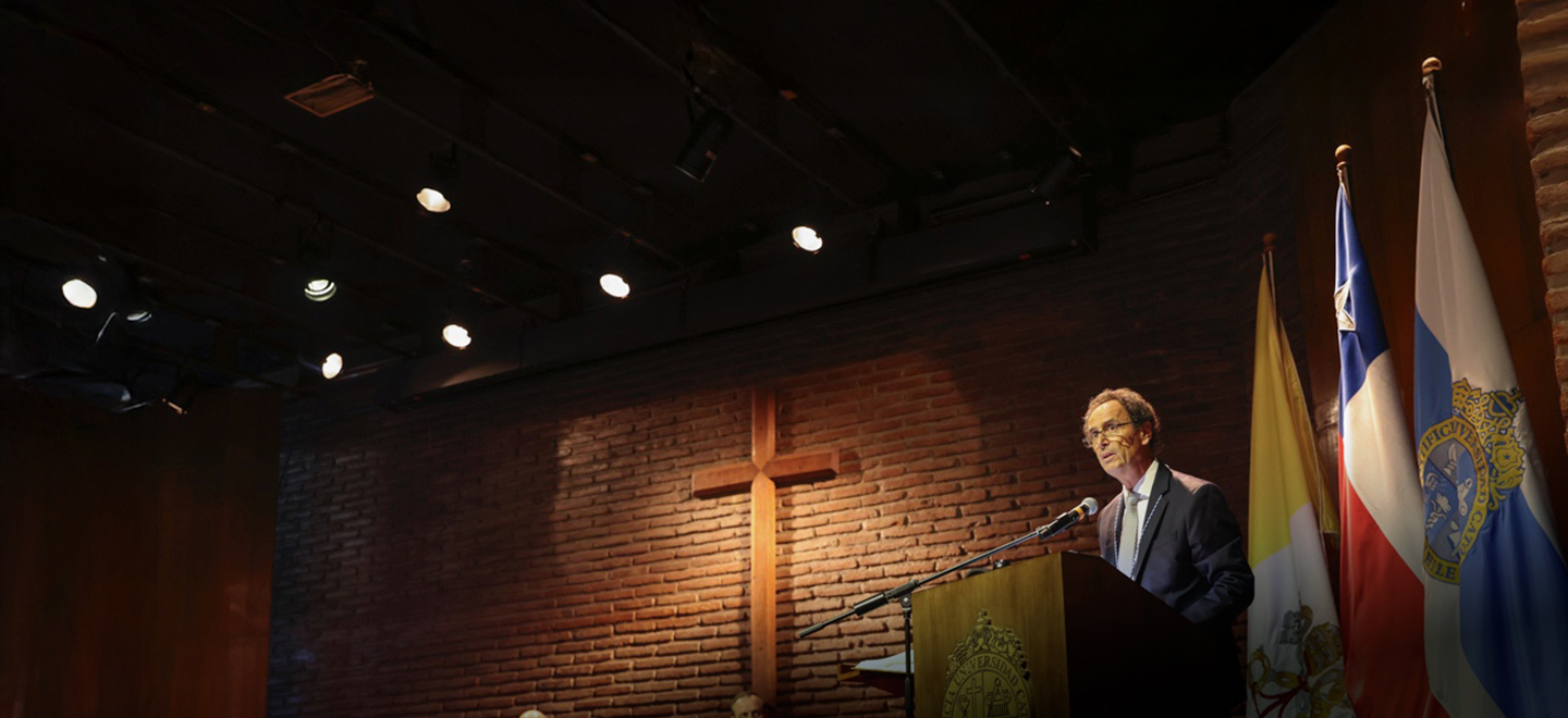 A man in a dark suit and tie speaks at a wooden podium in a dimly lit auditorium. A large wooden cross is mounted on a brick wall behind him. Flags, including those of Chile and the Vatican, are displayed on one side of the stage.