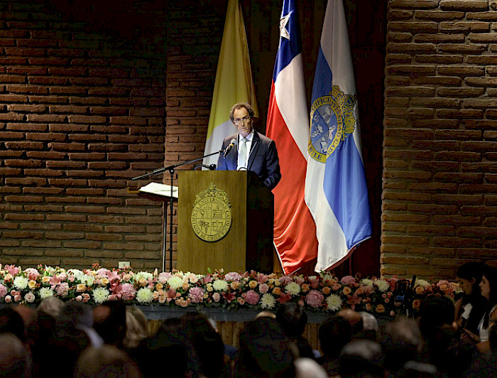 A man in a dark suit and tie speaks at a wooden podium in a dimly lit auditorium. A large wooden cross is mounted on a brick wall behind him. Flags, including those of Chile and the Vatican, are displayed on one side of the stage.